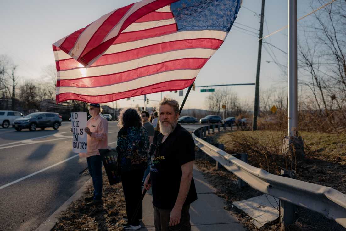 William Angus, 55 ans, résident du comté de Warren, au centre, participe à la protestation contre l'achat par les services d'immigration et de douane des États-Unis d'un entrepôt au 1879 route 46 qui doit être utilisé comme installation de traitement des immigrants sur Ledgewood Avenue et route 183 à Netcong, canton de Roxbury, New Jersey, États-Unis, le mardi 10 mars 2026. CRÉDIT : José A. Alvarado Jr. pour NPR, @josealvarado