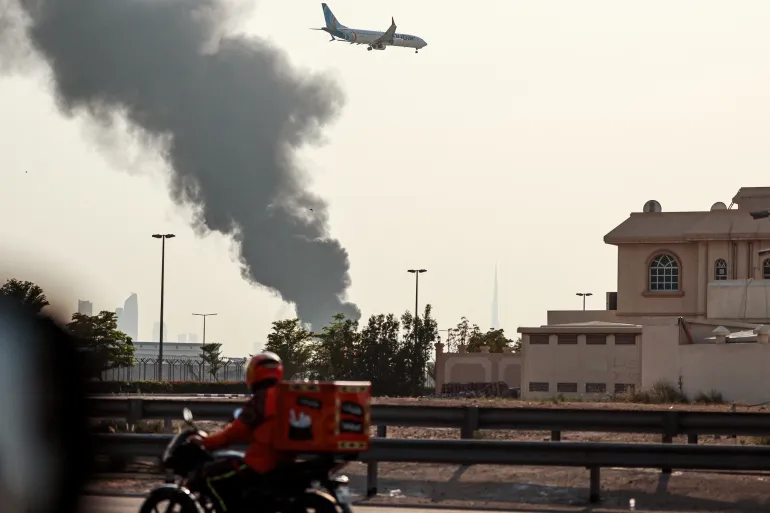 Un vélo de livraison roule le long d'une autoroute alors qu'un avion international FlyDubai sur la côte basse se prépare à atterrir alors qu'un panache de fumée s'élève d'un incendie en cours près de l'aéroport international de Dubaï, à Dubaï, le 16 mars 2026.