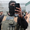 Un agent de l’Immigration et des Douanes, portant un masque tactique et un gilet, tient un téléphone pour filmer une vidéo.