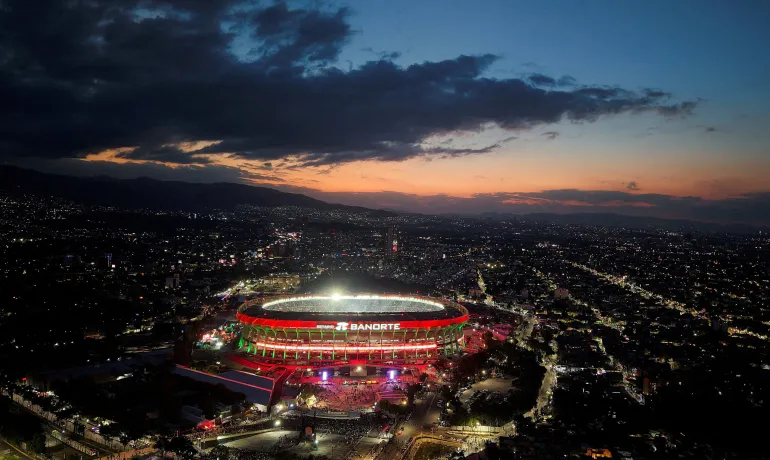 Le stade Azteca s'illumine au coucher du soleil, avec une ville sombre derrière et des nuages ​​au-dessus