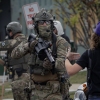 Un agent des forces de l’ordre fédérales vêtu de vêtements de camouflage affronte une manifestante portant une casquette de baseball bleue, dos à la caméra.
