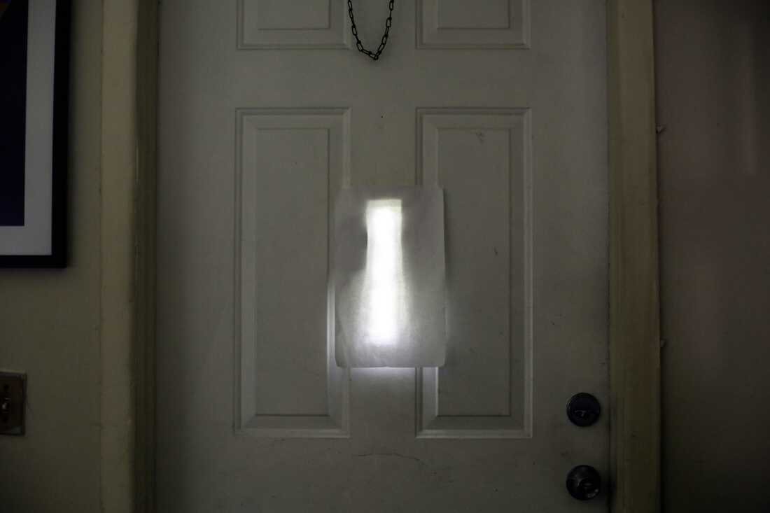 Une feuille de papier blanc recouvre une petite fenêtre étroite au milieu de la porte d'entrée blanche de la maison de Farah Larrieux, photographiée de l'intérieur.