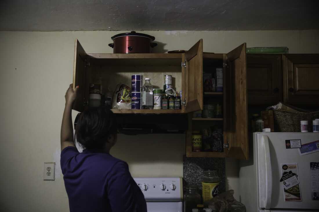 Photographiée dos à la caméra, Farah Larrieux ouvre un meuble de cuisine situé au-dessus d'une cuisinière. L'armoire est remplie d'agrafes de garde-manger.